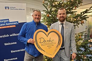 1. Vorstand Marco Heuschmann zusammen mit einem Vertreter der Bank bei der Spendenübergabe, ein "Danke"-Schild in Herzform haltend, im Hintergrund ein Weihnachtsbaum